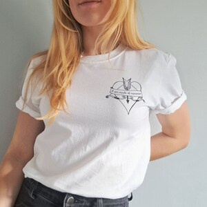 Puede incluir: Camiseta blanca con mangas enrolladas. Un gr&aacute;fico negro en forma de coraz&oacute;n con una flecha y una pancarta que dice "I am made of memories" est&aacute; impreso en el pecho. La camiseta est&aacute; hecha de un material suave.