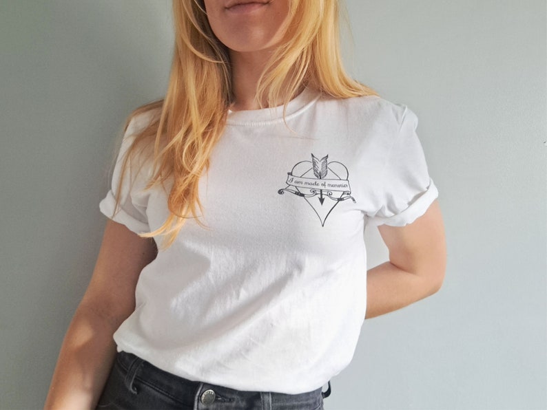 Puede incluir: Camiseta blanca con mangas enrolladas. Un gr&aacute;fico negro en forma de coraz&oacute;n con una flecha y una pancarta que dice "I am made of memories" est&aacute; impreso en el pecho. La camiseta est&aacute; hecha de un material suave.
