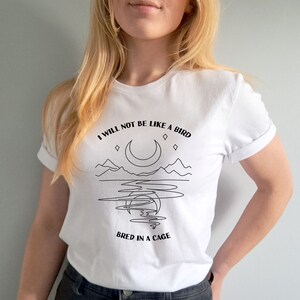 May include: White t-shirt with a black line drawing of a crescent moon, mountains, and water. The text "I will not be like a bird bred in a cage" is printed on the shirt.