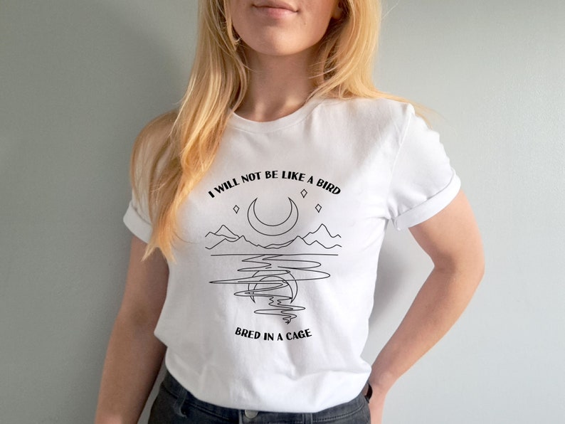 May include: White t-shirt with a black line drawing of a crescent moon, mountains, and water. The text "I will not be like a bird bred in a cage" is printed on the shirt.