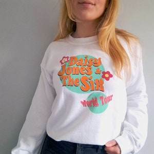 Op de afbeelding: Witte trui met een grafische print van "Daisy Jones & The Six World Tour" in een retro stijl met een kleurenschema in groen, oranje en roze.