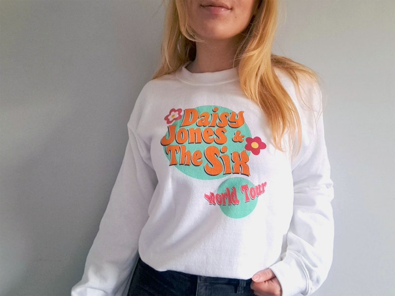 Op de afbeelding: Witte trui met een grafische print van "Daisy Jones & The Six World Tour" in een retro stijl met een kleurenschema in groen, oranje en roze.