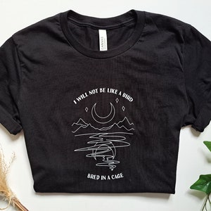 May include: Black t-shirt with a white graphic design featuring a crescent moon, mountains, and the text "I will not be like a bird bred in a cage."