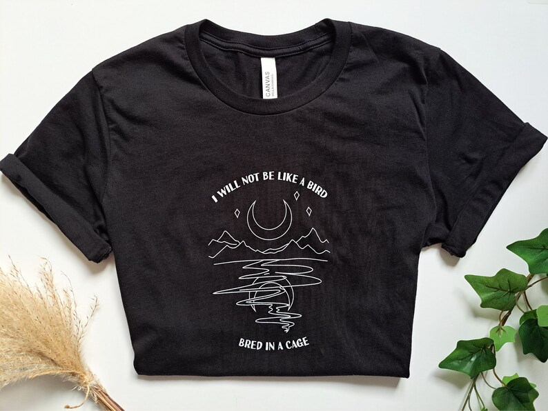 May include: Black t-shirt with a white graphic design featuring a crescent moon, mountains, and the text "I will not be like a bird bred in a cage."