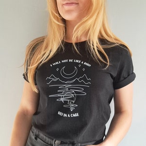 May include: Black t-shirt with a white graphic design featuring a crescent moon, mountains, and the text "I will not be like a bird caged in a cage."