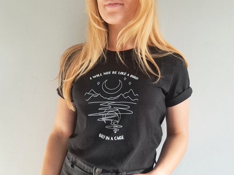 May include: Black t-shirt with a white graphic design featuring a crescent moon, mountains, and the text "I will not be like a bird caged in a cage."