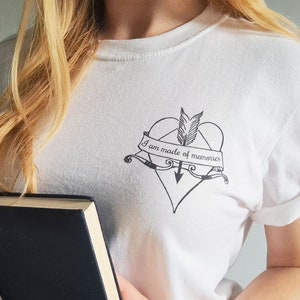 Puede incluir: Camiseta blanca con un gr&aacute;fico negro de un coraz&oacute;n atravesado por una flecha, con el texto "I am made of memories" en una pancarta. La camiseta tiene mangas enrolladas y cuello redondo. Un libro con una cubierta oscura se sostiene en la parte inferior de la imagen.