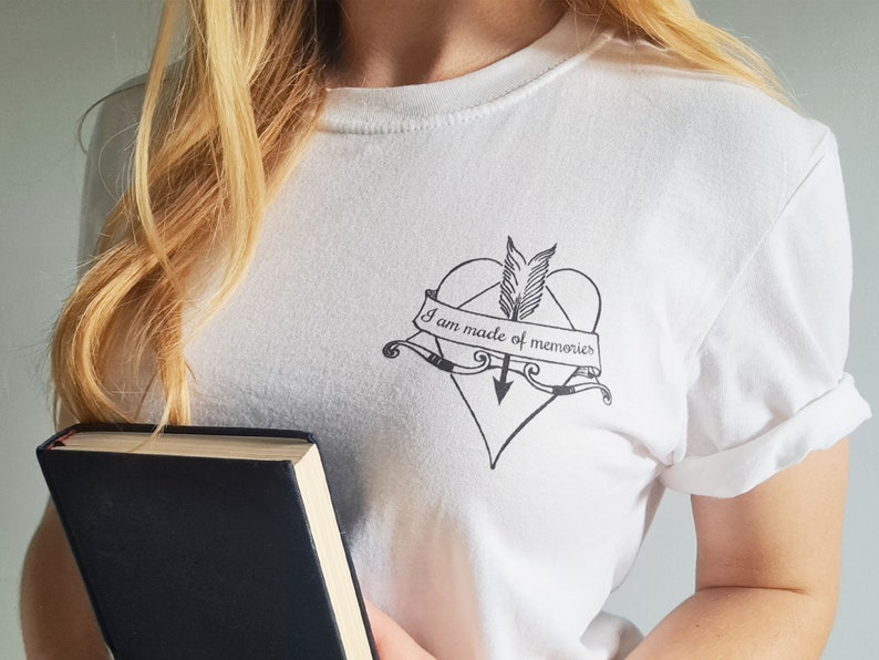 Puede incluir: Camiseta blanca con un gr&aacute;fico negro de un coraz&oacute;n atravesado por una flecha, con el texto "I am made of memories" en una pancarta. La camiseta tiene mangas enrolladas y cuello redondo. Un libro con una cubierta oscura se sostiene en la parte inferior de la imagen.