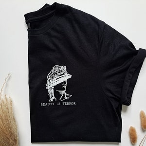 Może przedstawiać: Czarny T-shirt z białym nadrukiem przedstawiającym popiersie rzeźby z tekstem "BEAUTY IS TERROR" poniżej.