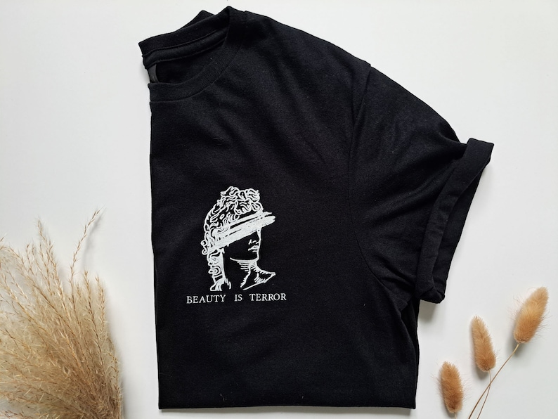 Może przedstawiać: Czarny T-shirt z białym nadrukiem przedstawiającym popiersie rzeźby z tekstem "BEAUTY IS TERROR" poniżej.