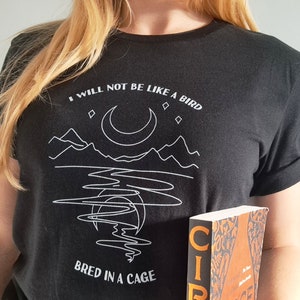 May include: A black t-shirt with a white graphic design of a crescent moon, mountains, and a body of water. The text "I will not be like a bird bred in a cage" is printed on the shirt.