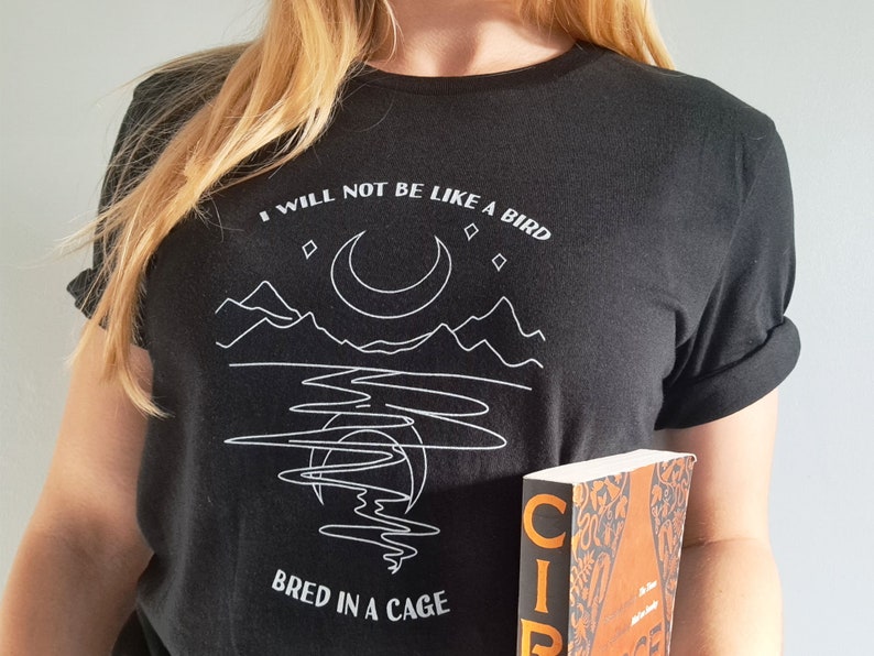 May include: A black t-shirt with a white graphic design of a crescent moon, mountains, and a body of water. The text "I will not be like a bird bred in a cage" is printed on the shirt.