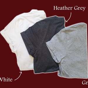 May include: Three t-shirts in different colours: white, heather grey, and grey. The t-shirts are laid out flat on a red background.