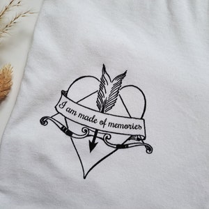 Puede incluir: Camiseta blanca con un coraz&oacute;n negro atravesado por una flecha, con un dise&ntilde;o de pluma y arco. Una pancarta en el coraz&oacute;n dice "I am made of memories". La camiseta est&aacute; hecha de una tela blanca suave.