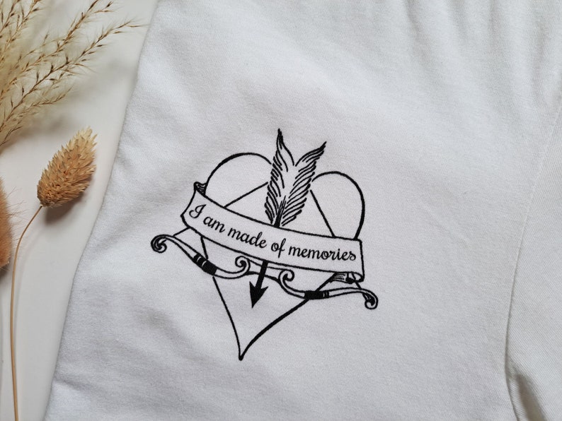 Puede incluir: Camiseta blanca con un coraz&oacute;n negro atravesado por una flecha, con un dise&ntilde;o de pluma y arco. Una pancarta en el coraz&oacute;n dice "I am made of memories". La camiseta est&aacute; hecha de una tela blanca suave.