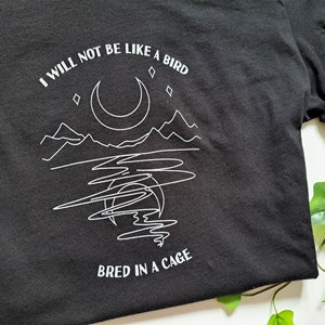 May include: Black t-shirt with a white graphic design of a moon, mountains, and a lake. The text "I will not be like a bird bred in a cage" is printed on the shirt.
