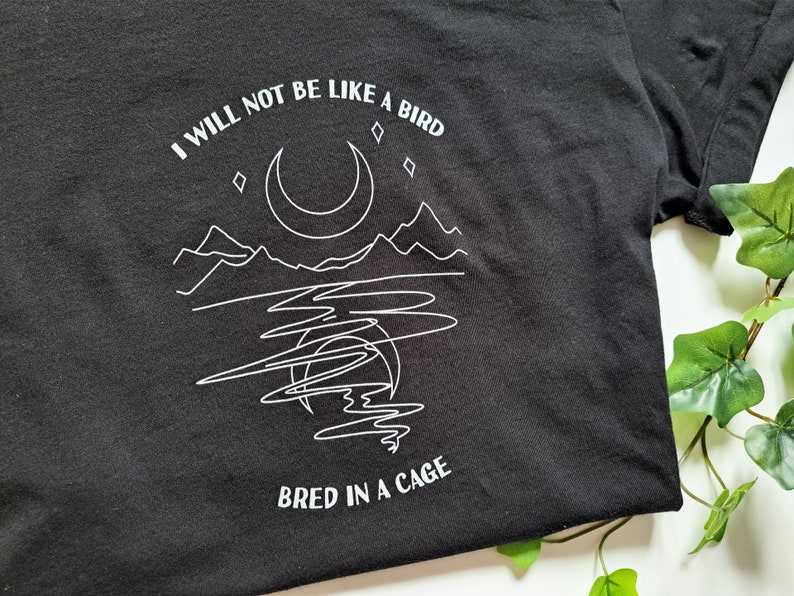 May include: Black t-shirt with a white graphic design of a moon, mountains, and a lake. The text "I will not be like a bird bred in a cage" is printed on the shirt.
