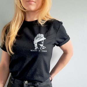 Może przedstawiać: Czarny t-shirt z białym nadrukiem przedstawiającym popiersie rzeźby z tekstem "BEAUTY IS TERROR" pod spodem.