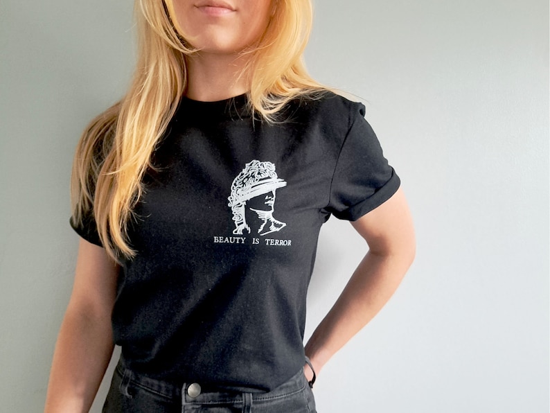 Może przedstawiać: Czarny t-shirt z białym nadrukiem przedstawiającym popiersie rzeźby z tekstem "BEAUTY IS TERROR" pod spodem.