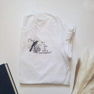 May include: White t-shirt featuring a black and white illustration of a bird with the text "It's a sin to kill a mockingbird."