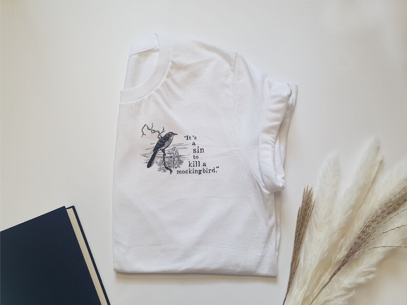 May include: White t-shirt featuring a black and white illustration of a bird with the text "It's a sin to kill a mockingbird."