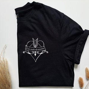 Puede incluir: Camiseta negra con un gr&aacute;fico blanco de un coraz&oacute;n atravesado por una flecha, plumas y el texto "I am made of memories". La camiseta est&aacute; doblada, mostrando la manga enrollada. Sobre una superficie blanca.