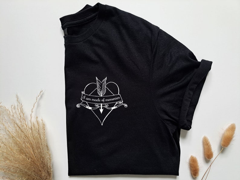 Puede incluir: Camiseta negra con un gr&aacute;fico blanco de un coraz&oacute;n atravesado por una flecha, plumas y el texto "I am made of memories". La camiseta est&aacute; doblada, mostrando la manga enrollada. Sobre una superficie blanca.
