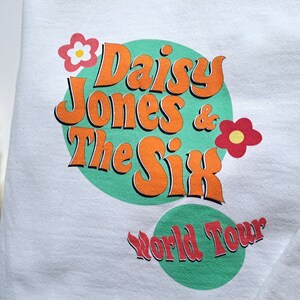 Op de afbeelding: Wit sweatshirt met een groene cirkel en oranje tekst die "Daisy Jones & The Six World Tour" luidt. De cirkel is versierd met twee rode en gele bloemen.