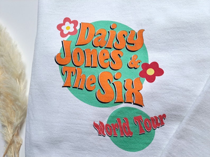 Op de afbeelding: Wit sweatshirt met een groene cirkel en oranje tekst die "Daisy Jones & The Six World Tour" luidt. De cirkel is versierd met twee rode en gele bloemen.