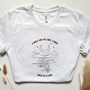 May include: White t-shirt with a black line drawing of a crescent moon, mountains, and a body of water. The text "I will not be like a bird bred in a cage" is printed below the image.
