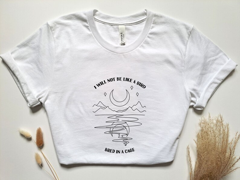 May include: White t-shirt with a black line drawing of a crescent moon, mountains, and a body of water. The text "I will not be like a bird bred in a cage" is printed below the image.