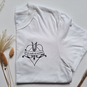 Puede incluir: Camiseta blanca con un dise&ntilde;o gr&aacute;fico negro. El dise&ntilde;o presenta un coraz&oacute;n atravesado por una flecha, una pluma y una pancarta con el texto "I am made of memories". La camiseta est&aacute; doblada, con el dise&ntilde;o visible en la parte delantera.