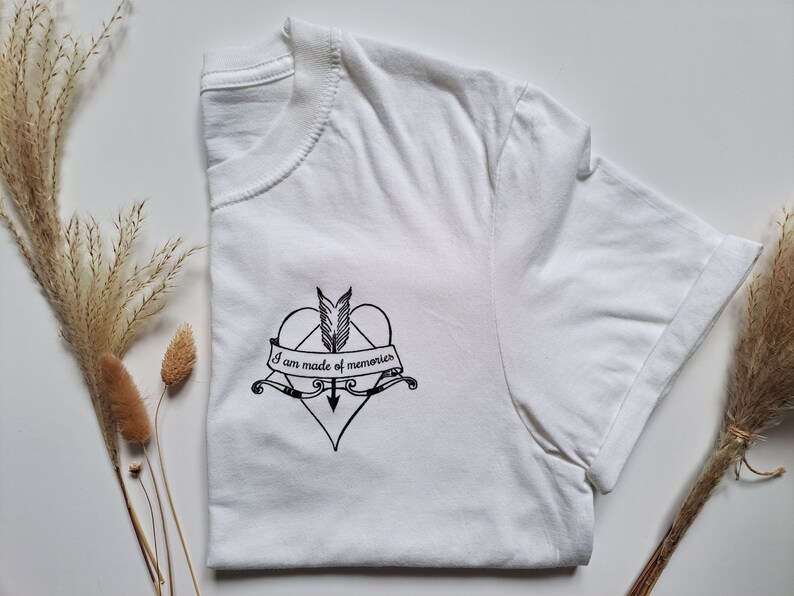 Puede incluir: Camiseta blanca con un dise&ntilde;o gr&aacute;fico negro. El dise&ntilde;o presenta un coraz&oacute;n atravesado por una flecha, una pluma y una pancarta con el texto "I am made of memories". La camiseta est&aacute; doblada, con el dise&ntilde;o visible en la parte delantera.
