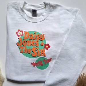 Op de afbeelding: Witte trui met een groene en oranje grafische print met de tekst "Daisy Jones & The Six World Tour" met een bloemendesign.