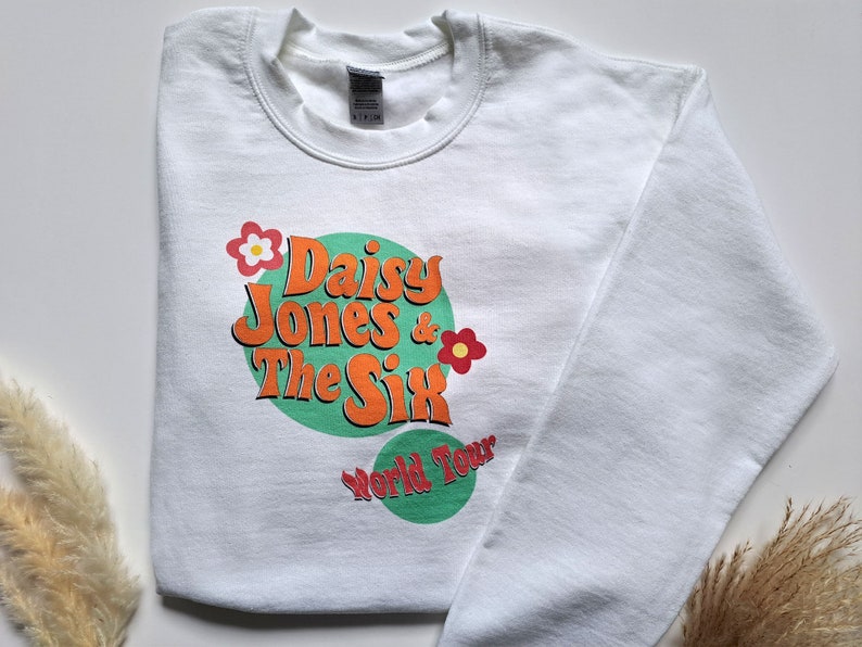 Op de afbeelding: Witte trui met een groene en oranje grafische print met de tekst "Daisy Jones & The Six World Tour" met een bloemendesign.