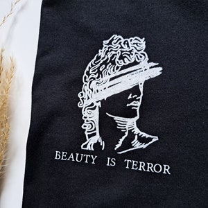 Może przedstawiać: Czarny T-shirt z białym nadrukiem przedstawiającym rzeźbę popiersia z zasłoniętymi oczami i tekstem "BEAUTY IS TERROR".