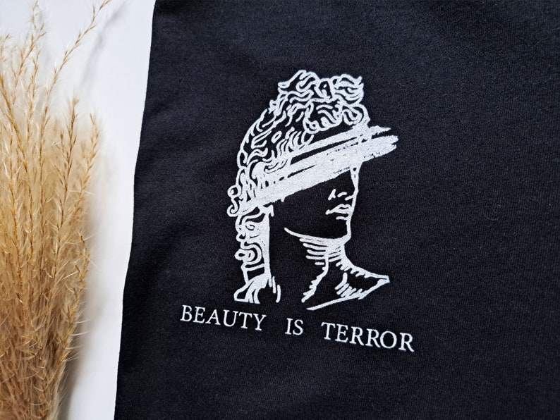 Może przedstawiać: Czarny T-shirt z białym nadrukiem przedstawiającym rzeźbę popiersia z zasłoniętymi oczami i tekstem "BEAUTY IS TERROR".