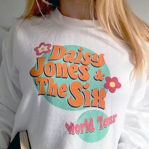 Op de afbeelding: Witte trui met een turquoise, oranje en roze grafische print met de tekst "Daisy Jones & The Six World Tour".