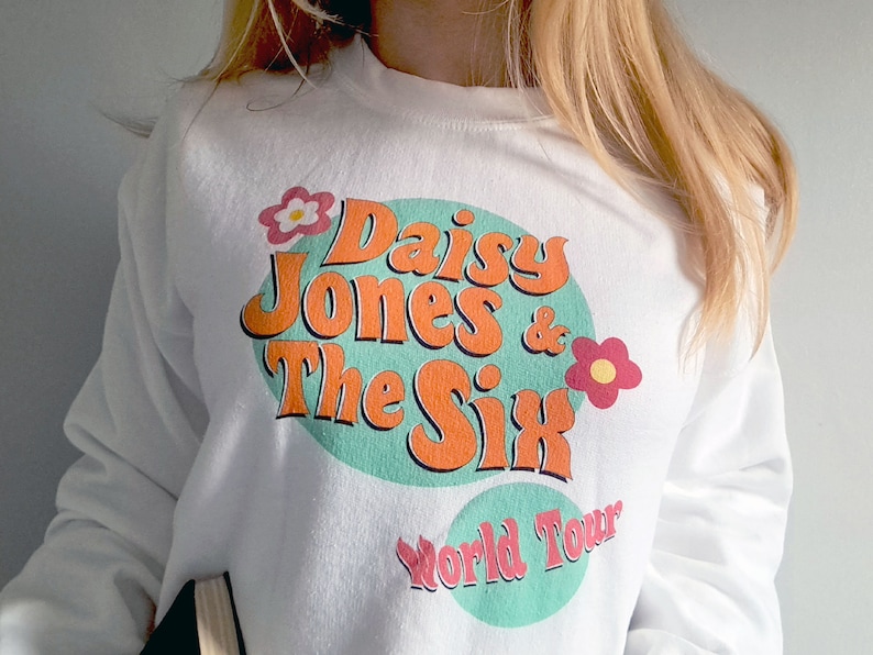 Op de afbeelding: Witte trui met een turquoise, oranje en roze grafische print met de tekst "Daisy Jones & The Six World Tour".