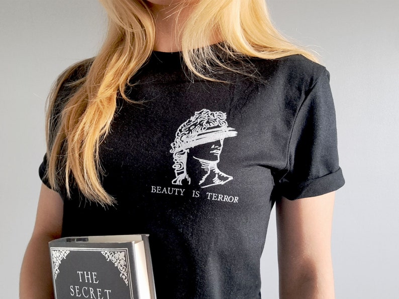 Może przedstawiać: Czarny T-shirt z białym nadrukiem przedstawiającym popiersie z koroną i tekstem "BEAUTY IS TERROR". Osoba trzyma czarną książkę z tekstem "THE SECRET" na okładce.