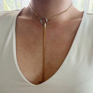Può includere: Una collana a catena dorata con un ciondolo rotondo. La collana è a strati, con una catena più corta nella parte superiore e una catena più lunga che si estende verso il basso sulla parte anteriore del collo.