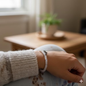 May include: A white beaded bracelet with a silver-colored charm is worn on a wrist. The bracelet is made of small, round beads. The charm has a decorative design. The person is wearing a gray sweater.