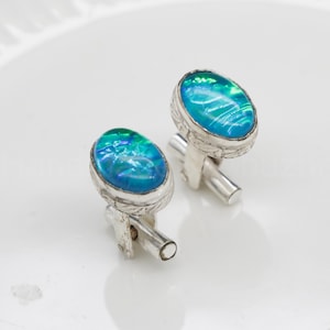 Puede incluir: Un par de gemelos de plata con piedras de ópalo ovaladas de color azul-verdoso iridiscente. Las piedras están engastadas en un marco de plata texturizado y detallado. Los gemelos tienen un diseño clásico y elegante.