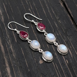 May include: Dangling earrings with a silver finish. Each earring features a teardrop-shaped, deep red gemstone at the top, followed by two oval, iridescent moonstone gems. The earrings are displayed against a dark wood grain background.