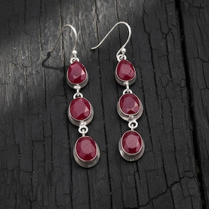 Puede incluir: Pendientes colgantes de plata con tres piedras preciosas ovaladas de color rojo intenso. Los pendientes están engastados en un bisel de plata y cuelgan de un gancho de plata. El fondo es una superficie oscura y texturizada.