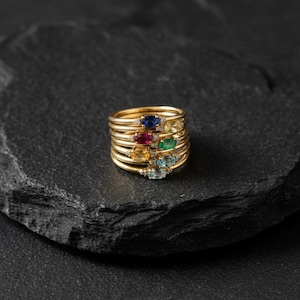 Peut inclure: Une pile de bagues en or ornées de pierres précieuses colorées : bleu, rouge, vert, jaune et bleu clair. Chaque bague, avec un fin anneau, est sertie d'une pierre ovale. Elles sont présentées sur une surface sombre et texturée.