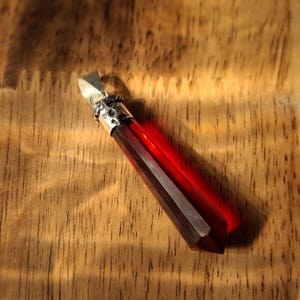 May include: A long, faceted, translucent red crystal pendant with a silver-toned metal cap featuring a decorative star pattern. The crystal has a pointed, hexagonal shape and is attached to a silver-toned bail for hanging on a necklace chain.