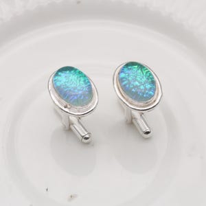 Puede incluir: Un par de gemelos plateados con centros ovalados iridiscentes verdes y azules. Los gemelos tienen un acabado pulido y un diseño clásico, adecuados para ocasiones formales o para añadir un toque de elegancia a cualquier atuendo.