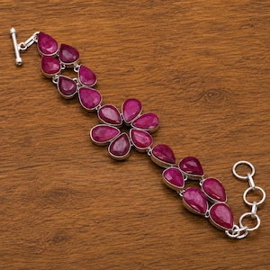 Pode incluir: Uma pulseira de prata com várias pedras preciosas rosa-escuras em forma de pera, dispostas num padrão floral ao centro, ladeadas por pedras adicionais ao longo da corrente, com um fecho de barra e uma série de elos circulares.