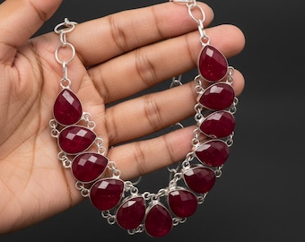 May include: A silver necklace featuring teardrop-shaped, faceted, deep red gemstones. The gemstones are set in a silver chain, creating a bold and elegant statement piece. The necklace is displayed against a dark background.